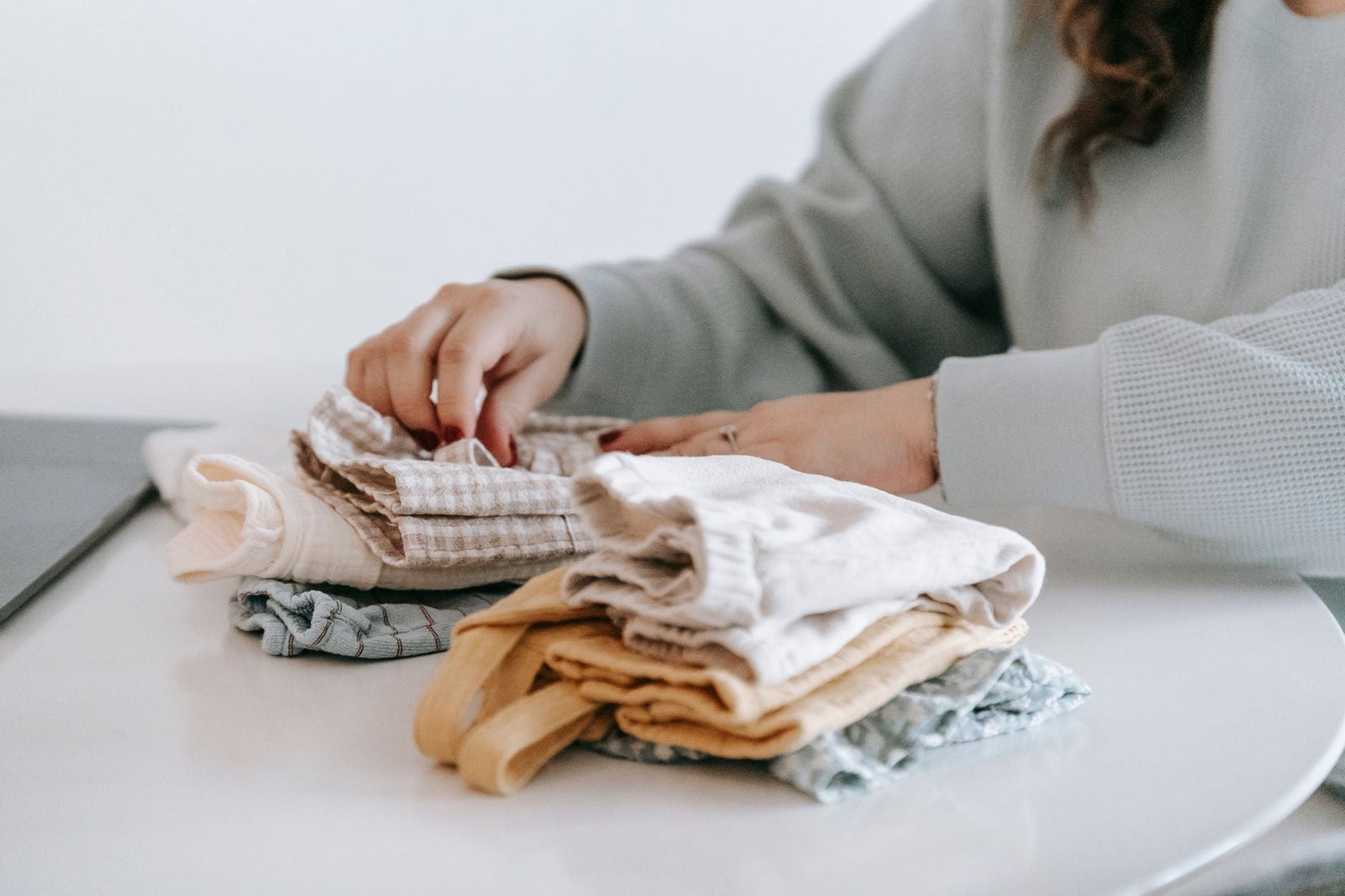 A woman's hands folding and arranging soft, neutral-toned baby muslins and garments on a white table.