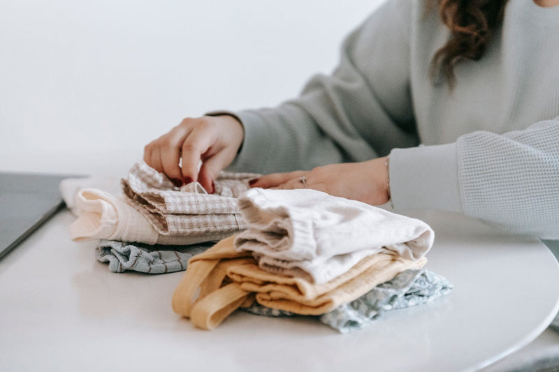 A woman's hands folding and arranging soft, neutral-toned baby muslins and garments on a white table.