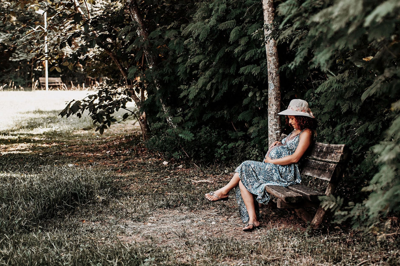 A pregnant woman in a sun hat sitting peacefully on a wooden bench in dappled tree shade.