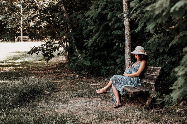 A pregnant woman in a sun hat sitting peacefully on a wooden bench in dappled tree shade.