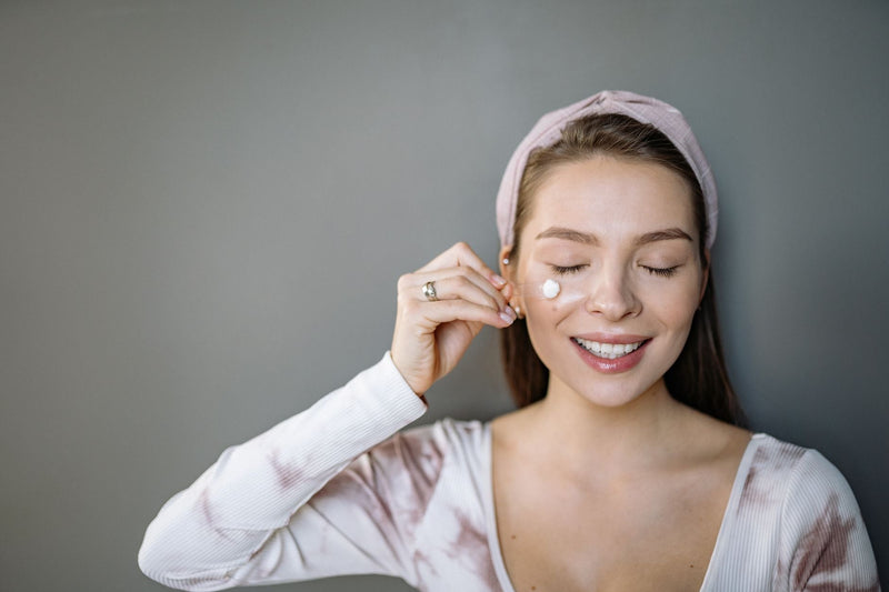 A smiling woman with a fabric headband gently applies moisturizer near her under-eye area against a plain gray background.