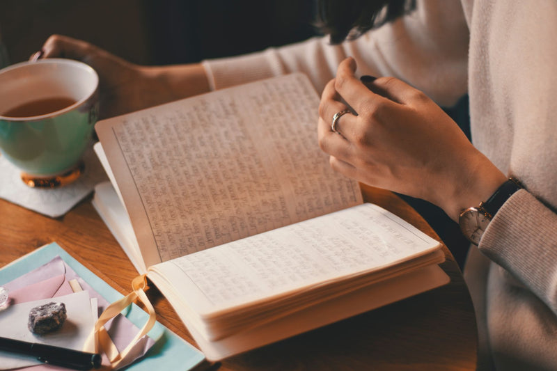 A woman's hands writing in a journal beside a cup of tea on a wooden table, a quiet, reflective morning moment.
