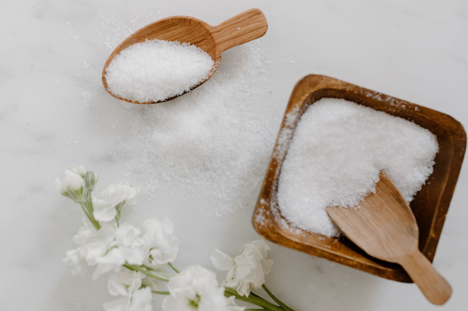 A minimalist flat lay of white bath salt in a wooden bowl with a wooden scoop and white flowers on a marble surface.