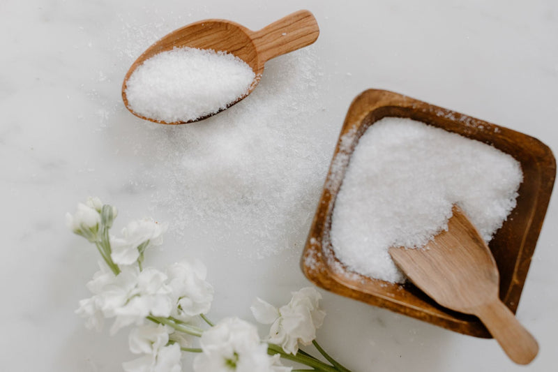 A minimalist flat lay of white bath salt in a wooden bowl with a wooden scoop and white flowers on a marble surface.