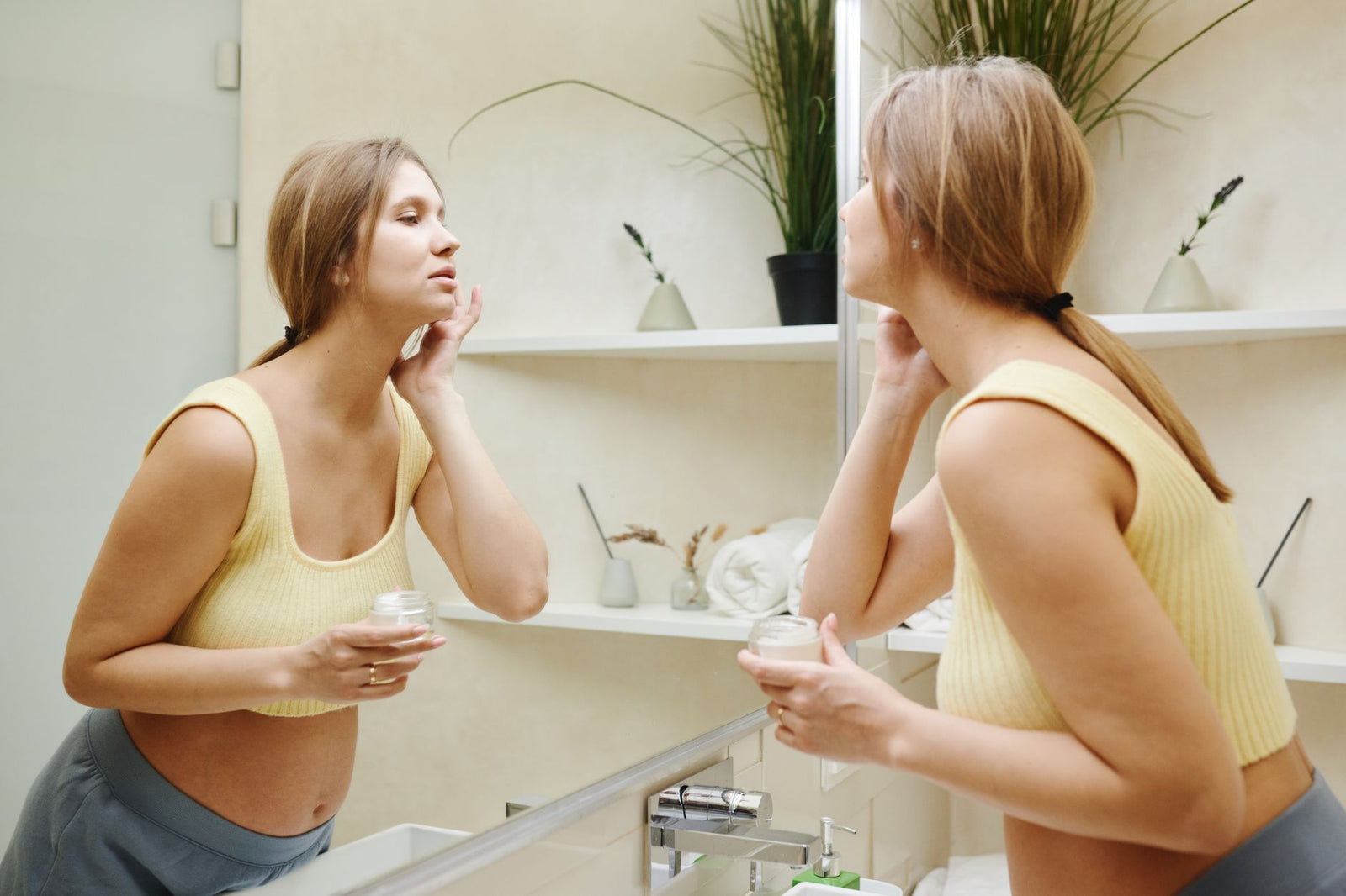A pregnant woman standing at a bathroom mirror, gently applying a skincare product to her face.