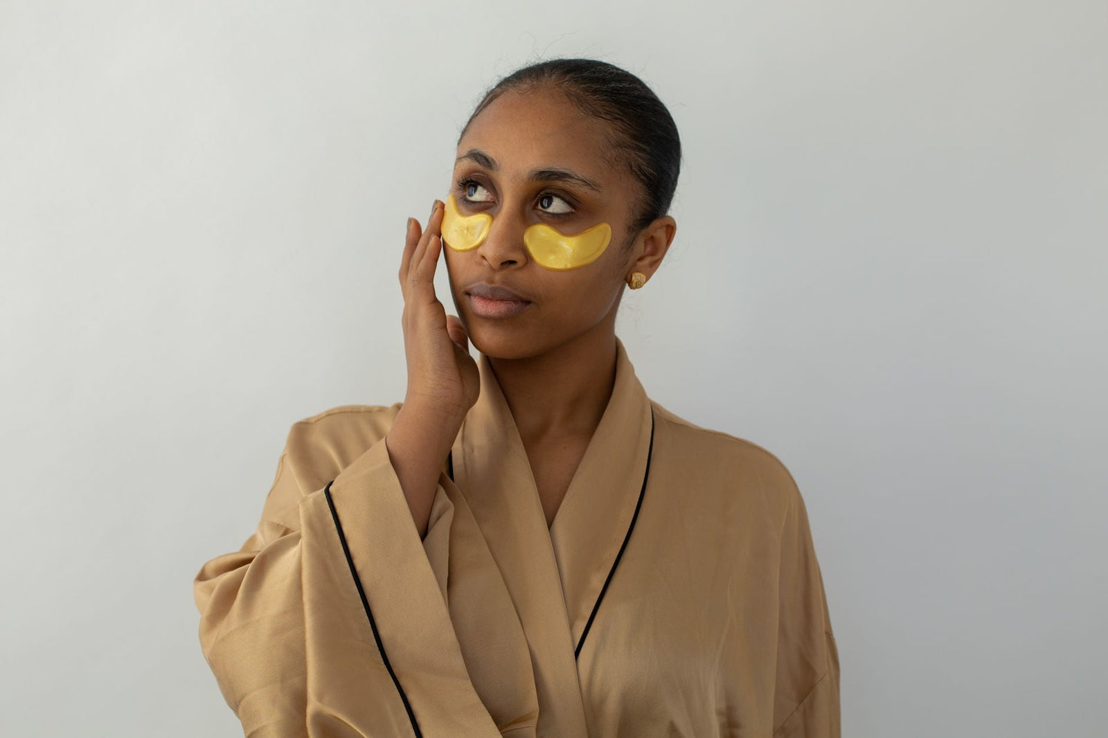 A woman in a silk robe wearing gold under-eye patches against a clean white background.