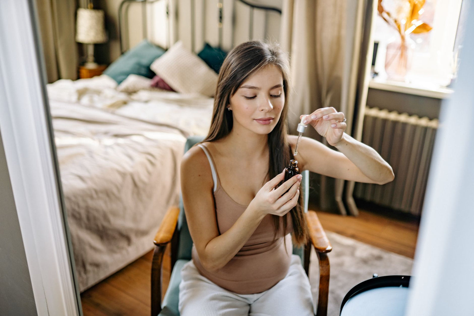 A pregnant woman seated in a bright bedroom, examining a dropper bottle of facial serum.