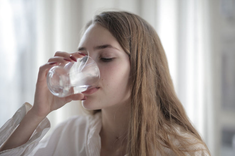 A woman drinking a glass of water at home, eyes closed, in soft natural light.