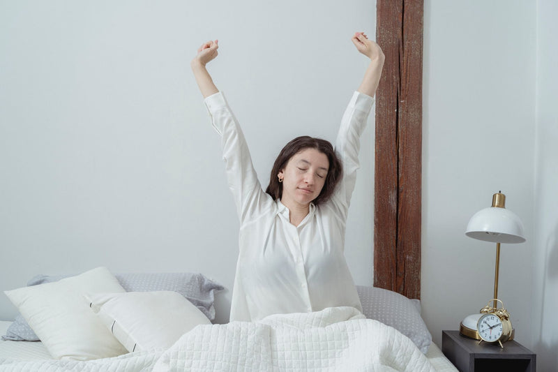 A woman with closed eyes calmly stretching her arms overhead while sitting up in a bright, white-linen bed in the morning.