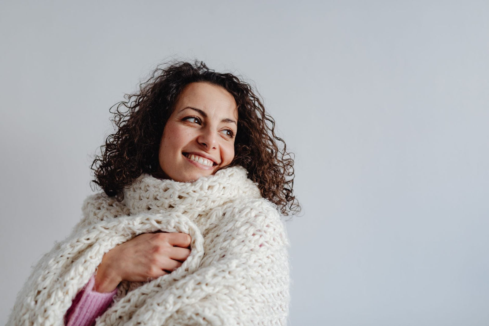 A woman wrapped snugly in a cream knit wrap, smiling warmly indoors.