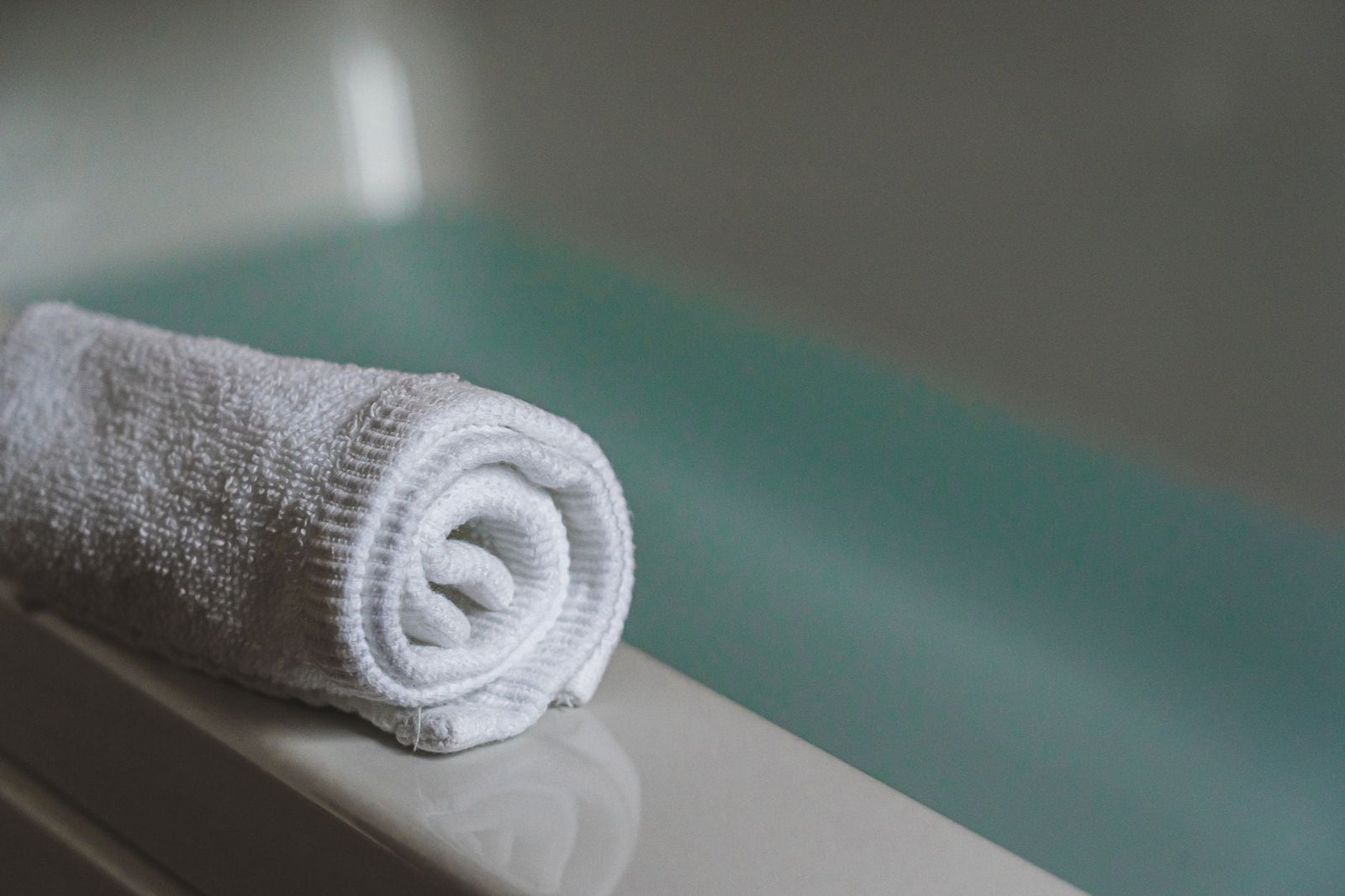 A rolled white towel resting on the edge of a tub, with soft teal water blurred in the background.