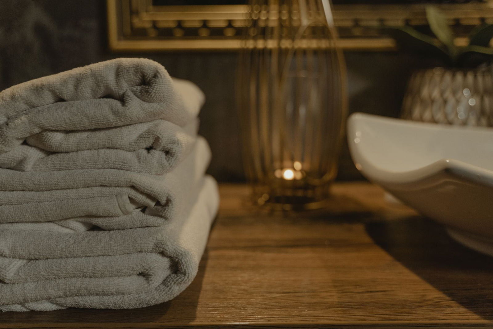 Neatly folded white towels beside a lit candle on a warm, softly lit bathroom shelf.