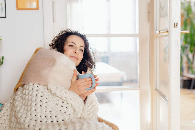 A woman wrapped in a cozy blanket holds a warm mug and looks out a sunlit window in a quiet, unhurried moment at home.