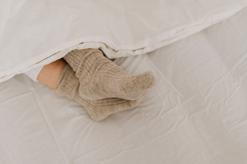 Comfortable feet in soft woolen socks peeking out from under a white linen blanket while resting on a bed.