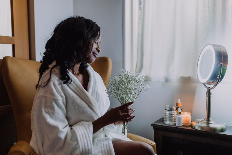 A woman in a soft white robe sits in a chair holding a small bunch of baby's breath flowers, beside a softly lit vanity mirror and skincare bottles on a nightstand.