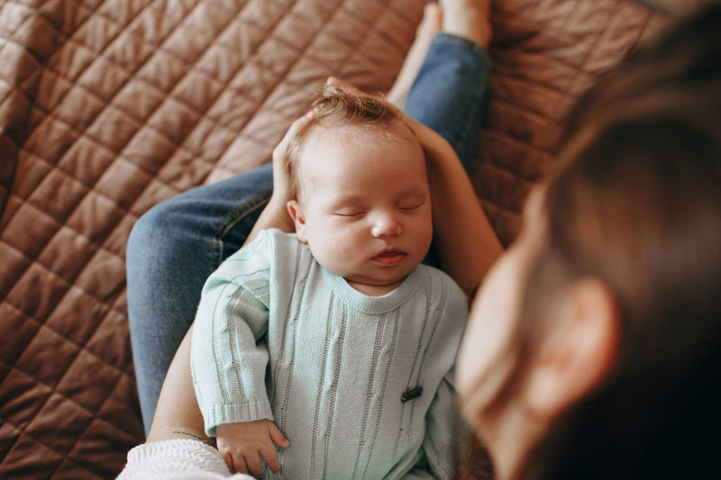 A newborn in a soft mint knit sleeps peacefully, cradled in a parent's lap on a quilted sofa.