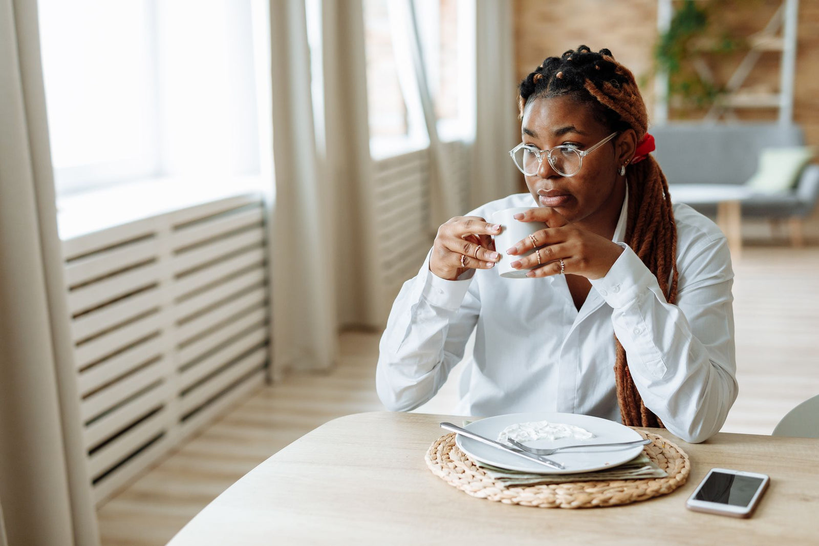A woman wearing clear-framed glasses sits calmly by a window, holding a cup of coffee at breakfast.