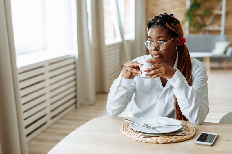 A woman wearing clear-framed glasses sits calmly by a window, holding a cup of coffee at breakfast.