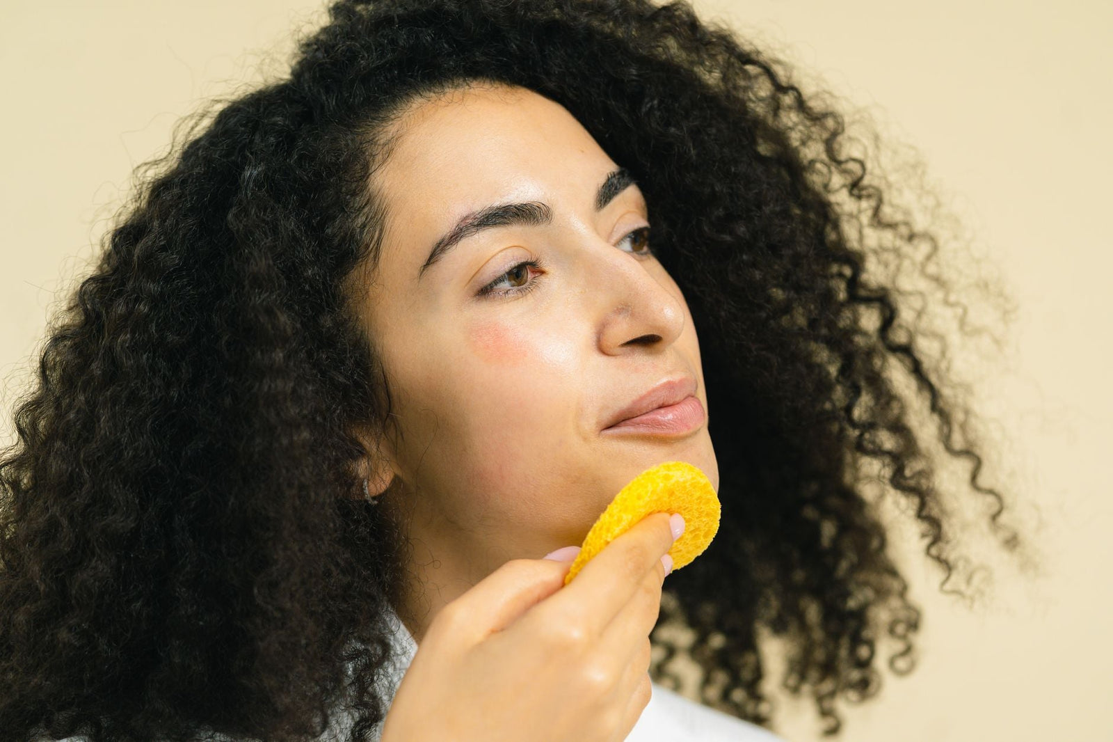 A woman gently pressing a soft yellow facial sponge against her cheek during a calm skincare routine.