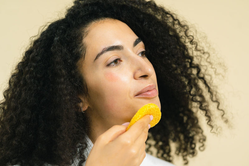 A woman gently pressing a soft yellow facial sponge against her cheek during a calm skincare routine.