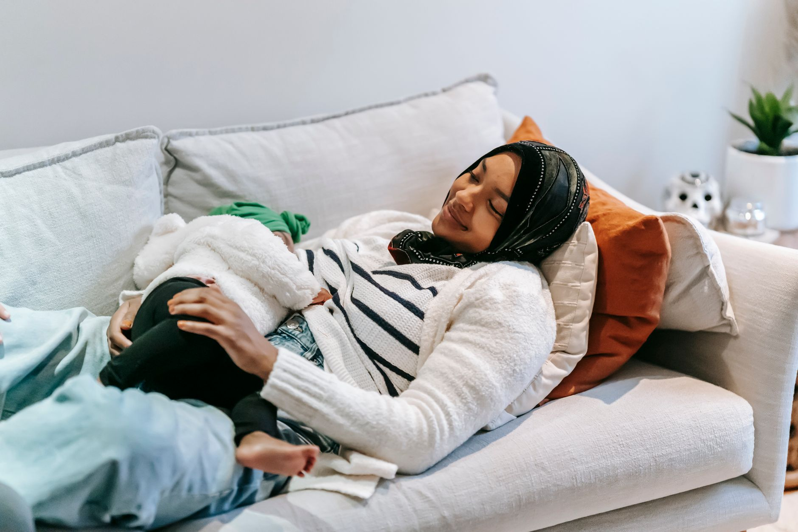 A smiling mother in a hijab breastfeeding her baby while reclining on a sofa at home.