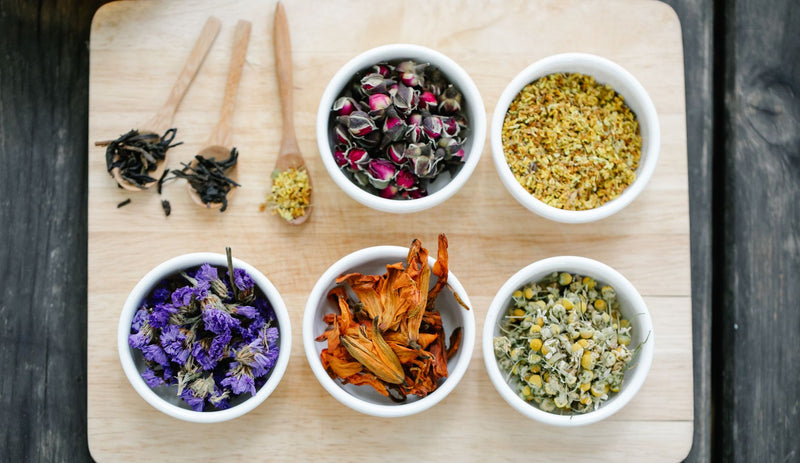 Small white bowls of dried calendula, chamomile, lavender, and rose petals with wooden spoons on a light wood board.